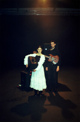 During the performance Masterpiece the performer Luisa Alfonso she is wearing a costume by Isabelle Lange, a white ruffled dress stands next to Peter Rubel, dressed in black, holding an electric guitar, in a dimly lit room with a spotlight. ISO LA - Isabelle Lange