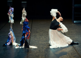The dancer Luisa Alfonso in a white ruffled skirt by Isabelle Lange, performs on a stage, gracefully arching back beside two tall, abstract sculptures adorned with colorful fabrics. ISO LA - Isabelle Lange