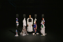 The dancer Luisa Alfonso stands on a dark stage flanked by large, colorful sculptures made of assorted fabrics, with four black speakers, designed by Isabelle Lange, positioned in the background. ISO LA - Isabelle Lange