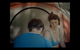 A scene from the movie Afterwater. Two people inside a tent, one with a blue shirt and short hair seen from the back, the other facing forward with a blue tank top and tattoos, appear in deep conversation. ISO LA - Isabelle Lange
