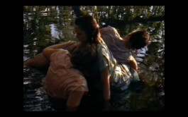 Three actors in wet dresses sit in a shallow pool of water, backlit by soft light filtering through trees, creating a serene and reflective mood. ISO LA - Isabelle Lange
