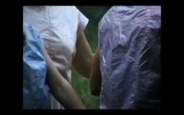 A scene from the movie Afterwater. Two people wearing rain ponchos, one blue and one purple, walking close together through a forested area. ISO LA - Isabelle Lange