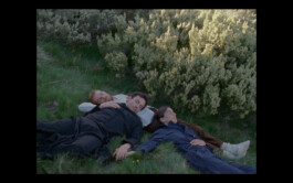 A scene from the movie Afterwater. Three people lying on the ground among bushy green shrubs, resting in a natural setting during twilight, appearing relaxed and contemplative. They are wearing black and blue overalls, a costume by Isabelle Lange ISO LA - Isabelle Lange
