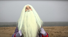 A person dressed in a fantasy costume by Isabelle Lange with a long, white beard and white hair, standing in a barren landscape with a foggy background. ISO LA - Isabelle Lange