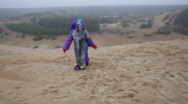 A person in a vibrant purple jacket and blue shawl stands at the edge of a sandy hill, overlooking a vast sandy landscape with sparse vegetation under a cloudy sky. ISO LA - Isabelle Lange