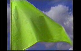 A bright green flag that is part of the textile installation path mark 07/22 by Isabelle Lange fluttering against a backdrop of a clear blue sky with sparse white clouds. ISO LA - Isabelle Lange