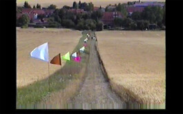 A line of colorful flags that are part of the textile installation path mark 07/22 by Isabelle Lange flutters along a narrow dirt path leading through a golden field towards distant houses under a dusky sky. ISO LA - Isabelle Lange