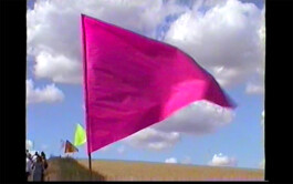 A bright pink flag that is part of the textile installation path mark 07/22 by Isabelle Lange fluttering against a backdrop of a clear blue sky with sparse white clouds, standing in what appears to be a sandy area, with other flags and people in the background. ISO LA - Isabelle Lange