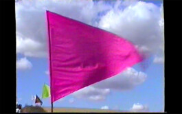 A bright pink flag that is part of the textile installation path mark 07/22 by Isabelle Lange fluttering against a backdrop of a clear blue sky with sparse white clouds, standing in what appears to be a sandy area, with other flags in the background. ISO LA - Isabelle Lange