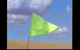A bright green flag that is part of the textile installation path mark 07/22 by Isabelle Lange fluttering against a backdrop of a clear blue sky with sparse white clouds, standing in what appears to be a sandy area. ISO LA - Isabelle Lange