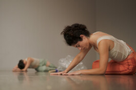 During the performance Tirana the performer Luisa Saraiva in a light tank top and orange pants, a costume by Isabelle Lange, focuses intently while stretching on a studio floor, another individual visible in the background. ISO LA - Isabelle Lange