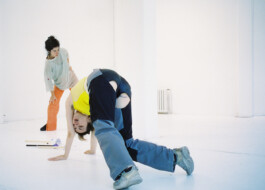 During the Performance Tirana a dancer performs a headstand in a brightly lit studio while another watches. they are wearing casual, colorful costumes by Isabelle Lange. ISO LA - Isabelle Lange