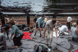 During the performance Tributary people move under a large, translucent and reflective tarp during an outdoor event, with some sitting on blankets and others walking or standing, they are wearing costumes by Isabelle Lange. ISO LA - Isabelle Lange