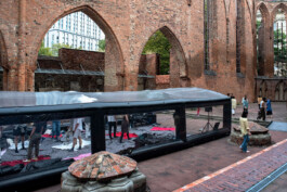 Exposed ruins in a glass enclosure at the base of a ruined church with arched brick walls, where several people are engaging in the performance Tributary. ISO LA - Isabelle Lange