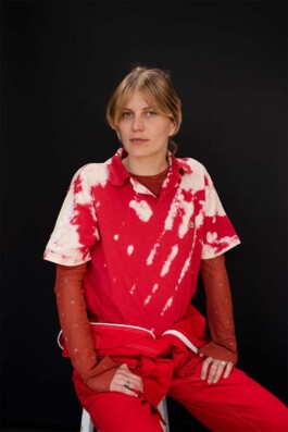 The costume designer and tailor Isabelle Lange sits on a stool against a black background, wearing a red tie-dye shirt and red pants. ISO LA - Isabelle Lange