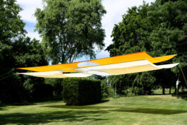 The textile Installation BALDACHINE by Isabelle Lange and Jakob Eichhorn, a large yellow and white canopy stretched over a sunny park area with lush green grass and a tall tree in the background under a clear blue sky. ISO LA - Isabelle Lange
