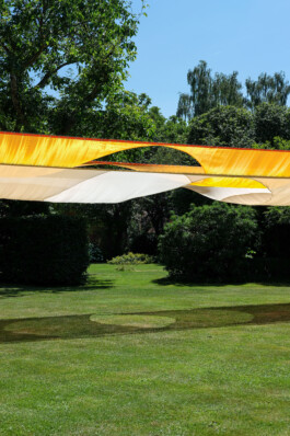 The textile installation BALDACHINE by Isabelle Lange and Jakob Eichhorn, which is a large yellow and beige canopy stretches over a lush green lawn in a garden, with dense trees and bright sunlight in the background. ISO LA - Isabelle Lange