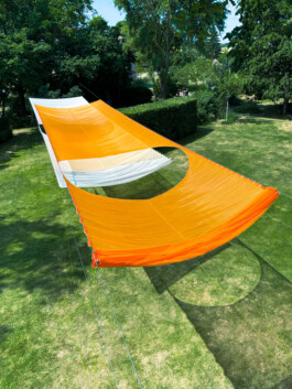 The textile Installation BALDACHINE by Isabelle Lange and Jakob Eichhorn, a large yellow and white canopy stretched over a sunny park area with lush green grass and a tall tree in the background under a clear blue sky, shot from birdseye perspective. ISO LA - Isabelle Lange